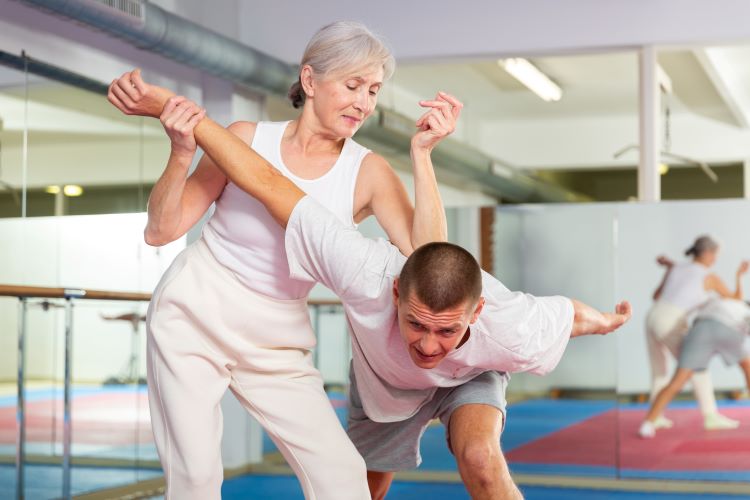 How To Defend Against Common Attacks for Senior Survivalists. Focused elderly woman performing elbow strike and wristlock, painful control move to immobilize male opponent during self defense training in gym.